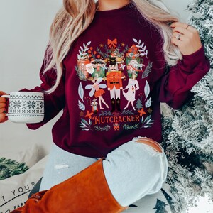 May include: A maroon sweatshirt with a festive illustration of the Nutcracker and other characters from the ballet. The design features a whimsical, colorful style with a white background. The text "The Nutcracker" is printed in white below the illustration.