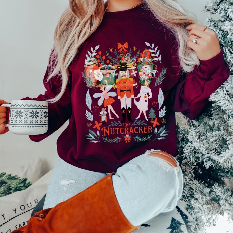 May include: A maroon sweatshirt with a festive illustration of the Nutcracker and other characters from the ballet. The design features a whimsical, colorful style with a white background. The text "The Nutcracker" is printed in white below the illustration.