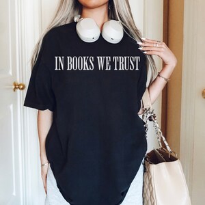 May include: A black oversized t-shirt with the text "IN BOOKS WE TRUST" printed in white on the front. The person is wearing grey sweatpants and has a beige handbag with a chain strap.