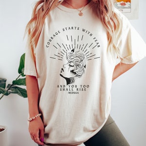 May include: A cream colored t-shirt with a black graphic of a woman with snakes for hair. The text on the shirt reads "Courage starts with fear and you too shall rise - Medusa".