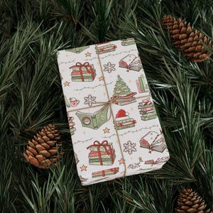 May include: A wrapped gift with a festive book-themed pattern. The white wrapping paper features illustrations of stacks of books, Christmas trees, snowflakes, and Santa hats. The gift is tied with twine and set on a bed of pine needles and pine cones.