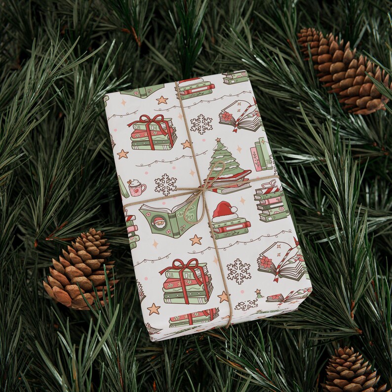 May include: A wrapped gift with a festive book-themed pattern. The white wrapping paper features illustrations of stacks of books, Christmas trees, snowflakes, and Santa hats. The gift is tied with twine and set on a bed of pine needles and pine cones.