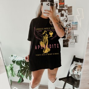 May include: Black t-shirt with a graphic print of a woman with wings holding a cup. The text on the shirt reads "Goddess World Tour", "5th Aug. 1998", "22:5682", "APHRODITE", and "Tickets Sold at Mount Olympus".
