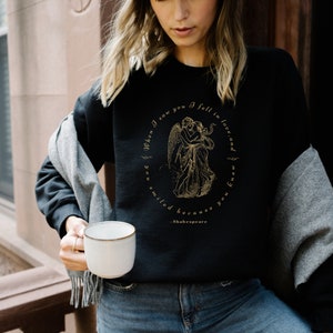 May include: Black sweatshirt with a gold graphic of two angels embracing. The graphic is surrounded by text that reads "When I saw you I fell in love, and you smiled because you knew" and "Shakespeare".