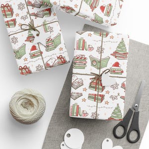 May include: Three wrapped gifts with a festive book-themed pattern. The wrapping paper features illustrations of books, Christmas trees, snowflakes, and Santa hats. Each gift is tied with twine and a blank tag. A ball of twine, scissors, and blank tags are also visible.