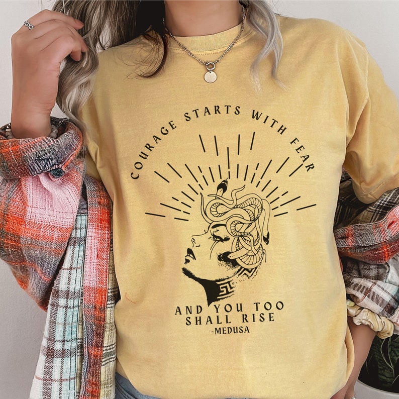 May include: A yellow t-shirt with a black graphic of a woman's face with snakes for hair. The text on the shirt reads "Courage starts with fear and you too shall rise - Medusa".