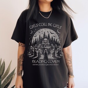 Może przedstawiać: Czarny t-shirt z graficznym wzorem przedstawiającym czaszkę, las i postacie w pelerynach. Tekst "GIRLS WILL BE GIRLS" i "READING COVEN" jest wydrukowany nad i pod grafiką, z dodatkowym tekstem na dole.