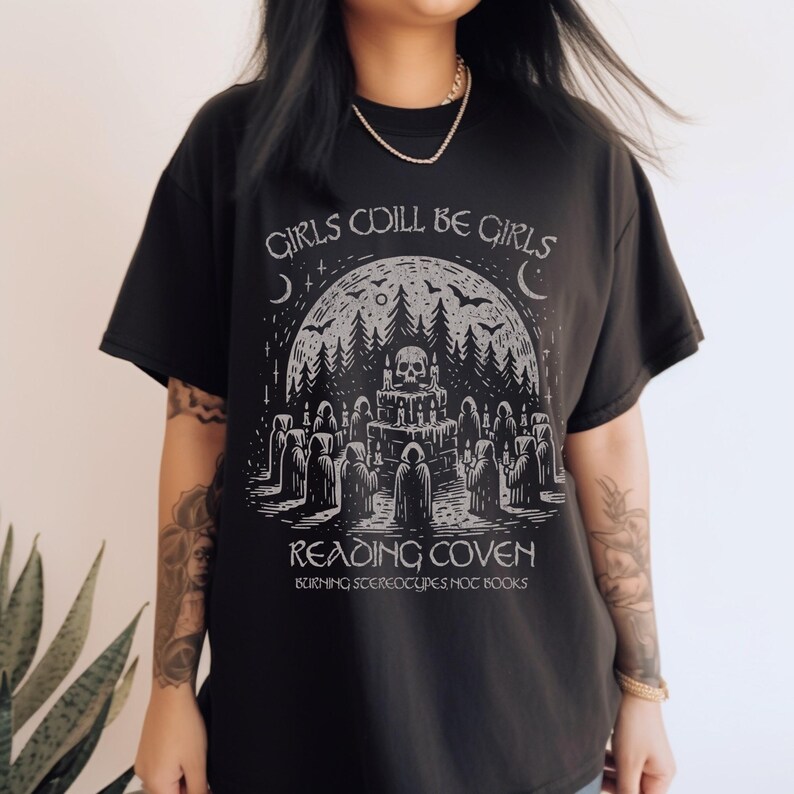 Może przedstawiać: Czarny t-shirt z graficznym wzorem przedstawiającym czaszkę, las i postacie w pelerynach. Tekst "GIRLS WILL BE GIRLS" i "READING COVEN" jest wydrukowany nad i pod grafiką, z dodatkowym tekstem na dole.