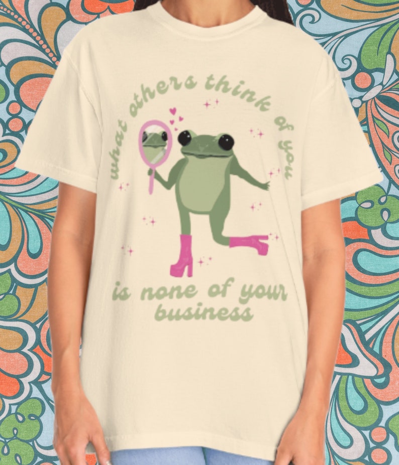May include: A light beige t-shirt with a green frog wearing pink boots and holding a pink mirror. The frog is looking at its reflection in the mirror. The text on the shirt reads "what others think of you is none of your business".