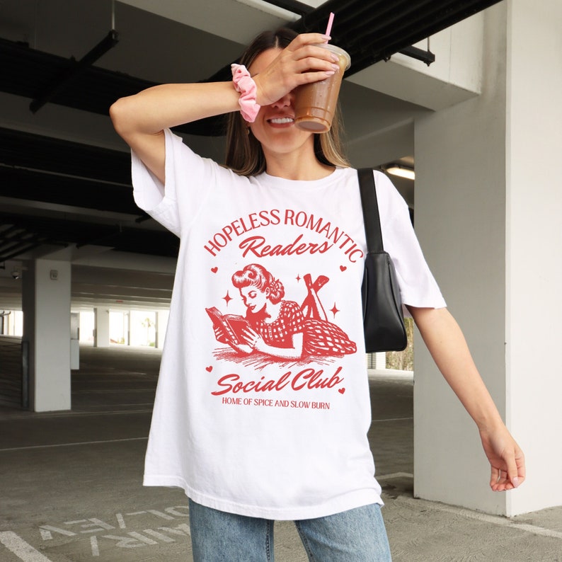 Peut inclure: T-shirt blanc avec un graphique rouge d'une femme lisant un livre. Le graphique indique "Hopeless Romantic Readers Social Club, Home of Spice and Slow Burn".