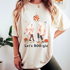 以下が含まれることがあります： クリーム色のTシャツに、2体の幽霊がディスコボールの下で踊っているグラフィックがプリントされています。幽霊の下には「Let's BOO-gie」というテキストがプリントされています。