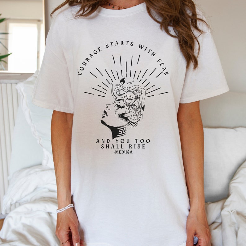 May include: White t-shirt with a black graphic of Medusa with snakes for hair. The text reads "Courage starts with fear and you too shall rise - Medusa".