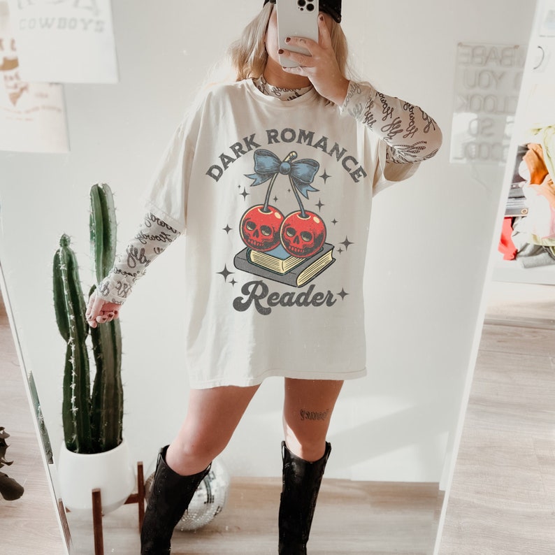 K&ouml;nnte beinhalten: Cremefarbenes &uuml;bergro&szlig;es T-Shirt mit dem Text "DARK ROMANCE READER" und einer Grafik von Kirschen mit Totenkopfgesichtern, einer blauen Schleife und einem Buch. Das Shirt wird mit einem gemusterten Langarmshirt und schwarzen Stiefeln getragen.
