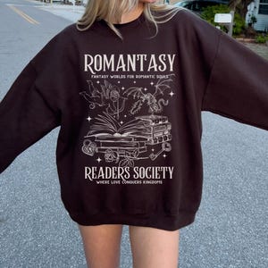 May include: A brown sweatshirt with a white graphic design. The graphic features a book, a dragon, an angel, and the text "Romantasy: Fantasy Worlds for Romantic Souls" and "Readers Society: Where Love Conquers Kingdoms".