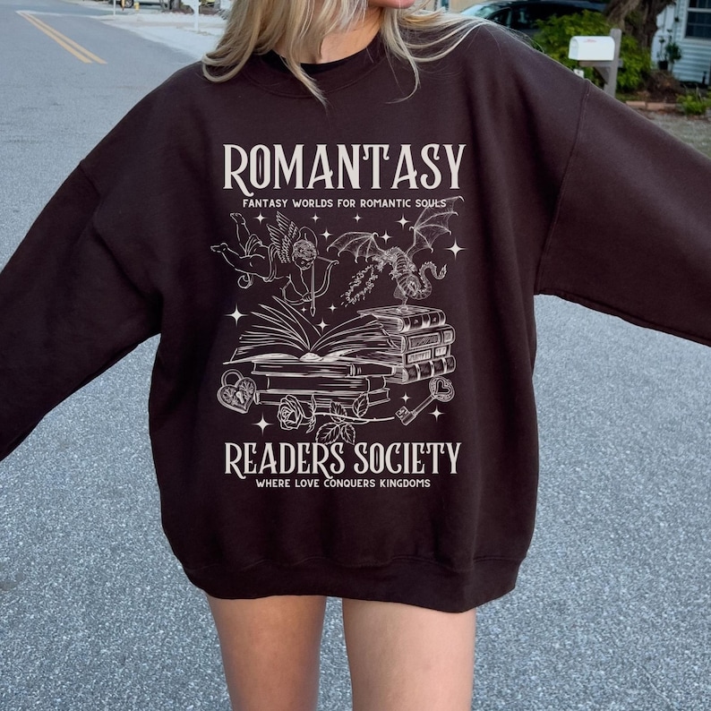 May include: A brown sweatshirt with a white graphic design. The graphic features a book, a dragon, an angel, and the text "Romantasy: Fantasy Worlds for Romantic Souls" and "Readers Society: Where Love Conquers Kingdoms".