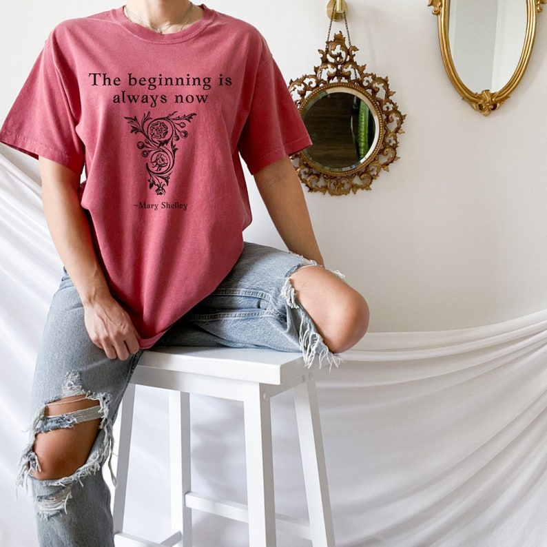 May include: A red t-shirt with a black floral design and the quote "The beginning is always now - Mary Shelley".