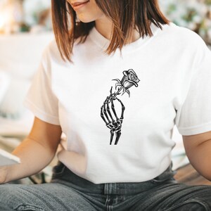 K&ouml;nnte beinhalten: Ein wei&szlig;es T-Shirt mit einem schwarzen Motiv einer Skeletthand, die eine Rose h&auml;lt.