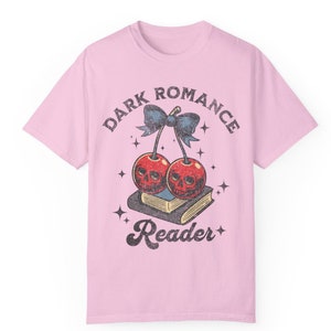 K&ouml;nnte beinhalten: Hellrosa T-Shirt mit einem Grafikdesign. Das Design zeigt zwei Kirschen mit Totenkopfgesichtern, eine blaue Schleife und die Worte "DARK ROMANCE Reader" in einem Vintage-Stil. Die Kirschen liegen auf einem Buch.