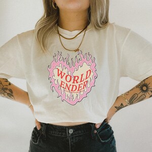 May include: A white t-shirt with a pink and yellow heart-shaped flame graphic. The text "WORLD ENDER" is written in red inside the heart. The text "YOUR MONSTERS YOUR TRUTH" is written in smaller red letters below the heart.