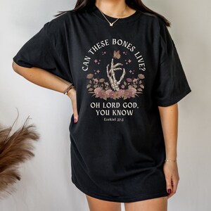 Puede incluir: Camiseta negra con un gr&aacute;fico de una mano esquel&eacute;tica sosteniendo una mariposa con el texto "Can these bones live? Oh Lord God, you know. Ezekiel 37:3"