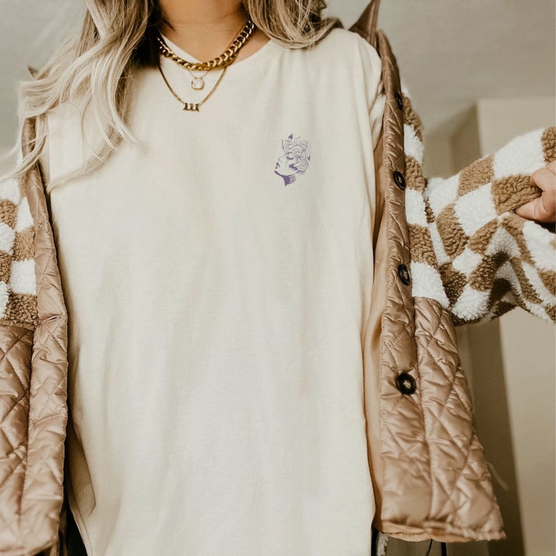 May include: A white long-sleeved t-shirt with a small, purple graphic of a woman's face on the left chest. The shirt is being worn by a person with long blonde hair and a gold necklace.
