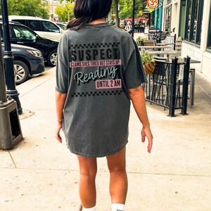 May include: A dark gray t-shirt with a black and white checkered pattern and the text "SUSPECT CLAIMS SHE'S TIRED BUT STAYS UP Reading UNTIL 2 AM" in black and pink lettering.