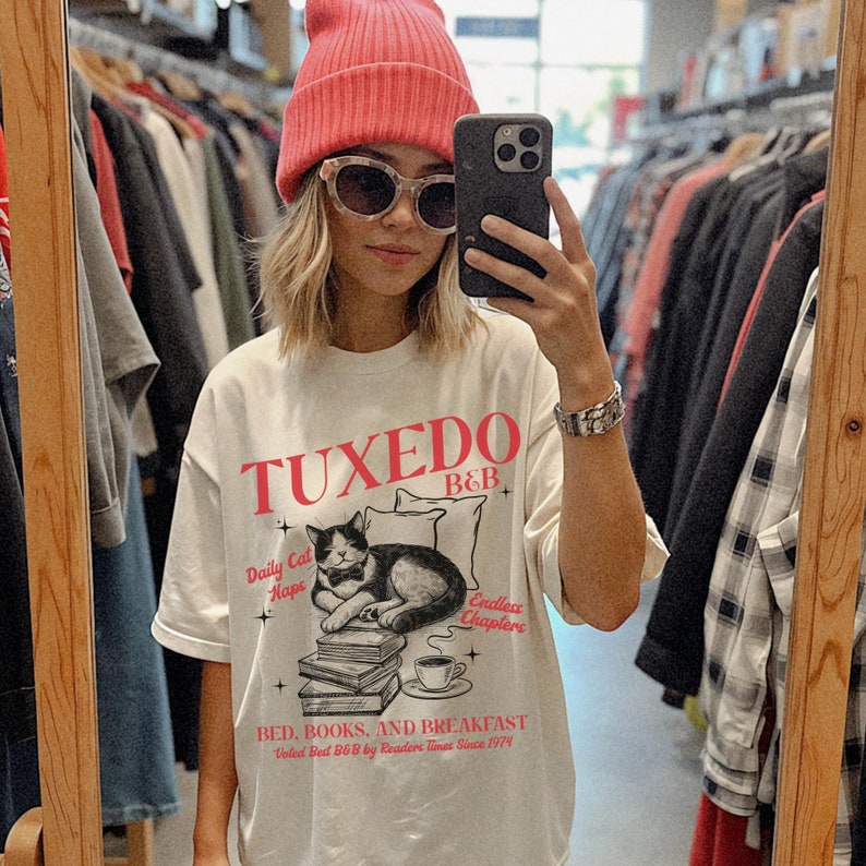 Op de afbeelding: Wit T-shirt met een zwart-witte illustratie van een kat die slaapt op een stapel boeken. Het shirt heeft rode tekst die "Tuxedo B&B" en "Daily Cat Naps" en "Endless Chapters" zegt. De tekst onderaan zegt "Bed, Books, and Breakfast" en "Voted Best B&B by Readers Times Since 1974".