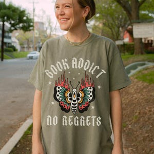 May include: Olive green t-shirt with a butterfly graphic and the text "BOOK ADDICT" above and "NO REGRETS" below. The butterfly has red, teal, and black details. The shirt is made of a soft material.