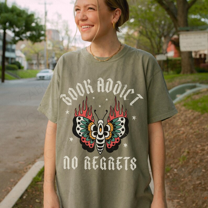 May include: Olive green t-shirt with a butterfly graphic and the text "BOOK ADDICT" above and "NO REGRETS" below. The butterfly has red, teal, and black details. The shirt is made of a soft material.