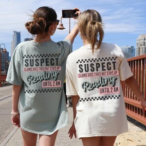 May include: Two people wearing light green and white t-shirts with the text "SUSPECT CLAIMS SHES TIRED BUT STAYS UP Reading UNTIL 2 AM" printed on the back. The shirts have a black and white checkered pattern.
