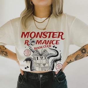 Pu&ograve; includere: T-shirt color crema con la scritta rossa "MONSTER ROMANCE BOOK CLUB". Il design include un'illustrazione di libri, cuori e tentacoli. La maglietta presenta anche le frasi "ONE BED, SIX ARMS, NO REGRETS, MEETS NIGHTLY" e "COME FOR THE PLOT, STAY FOR THE TENTACLES."