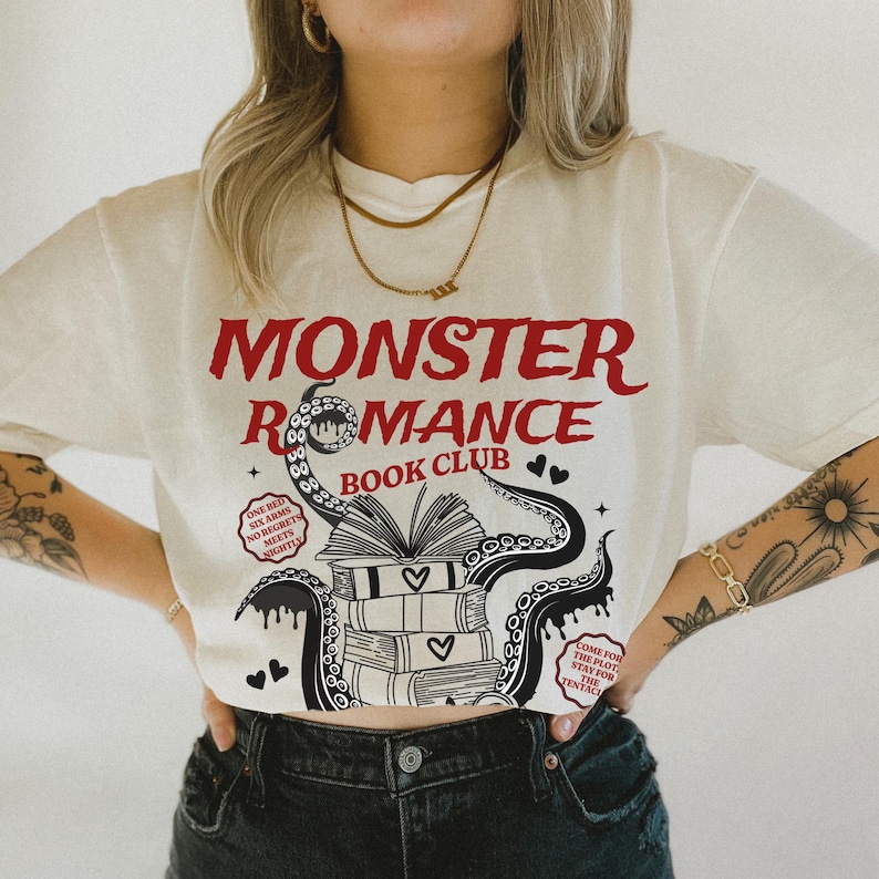 Pu&ograve; includere: T-shirt color crema con la scritta rossa "MONSTER ROMANCE BOOK CLUB". Il design include un'illustrazione di libri, cuori e tentacoli. La maglietta presenta anche le frasi "ONE BED, SIX ARMS, NO REGRETS, MEETS NIGHTLY" e "COME FOR THE PLOT, STAY FOR THE TENTACLES."