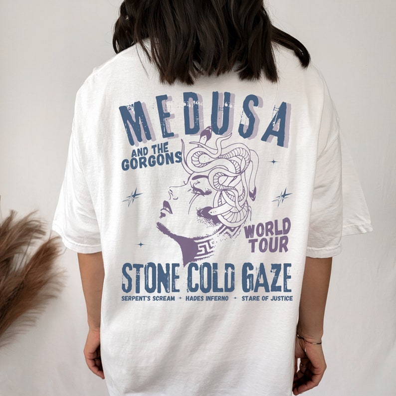 May include: White t-shirt with a graphic print of Medusa, a mythical Greek figure with snakes for hair. The text "MEDUSA AND THE GORGONS WORLD TOUR STONE COLD GAZE" is printed on the shirt. The text "SERPENT'S SCREAM HADES INFERNO STARE OF JUSTICE" is printed below the graphic.