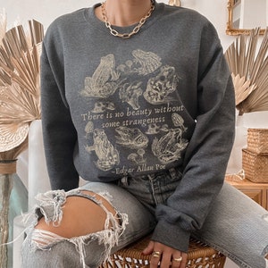 May include: A dark gray sweatshirt with a vintage illustration of frogs and mushrooms. The text "There is no beauty without some strangeness - Edgar Allan Poe" is printed on the front.