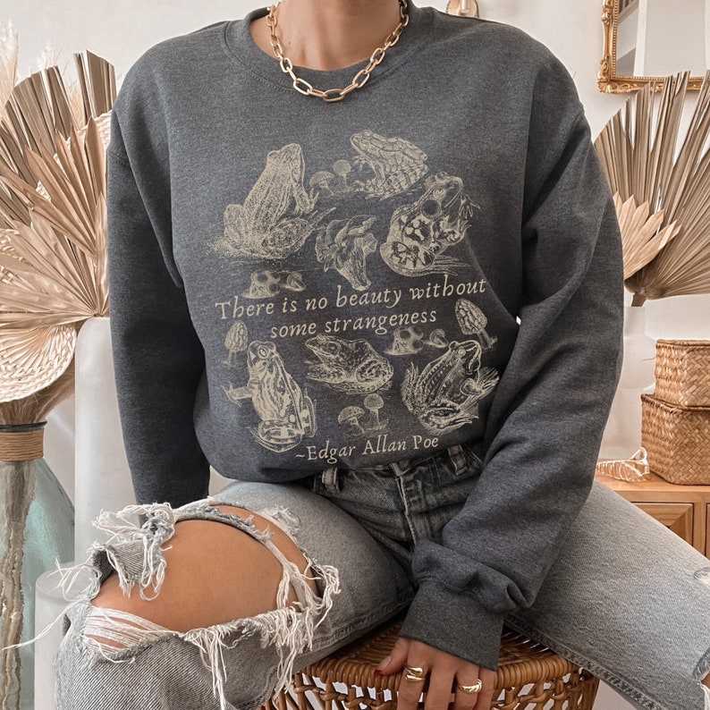 May include: A dark gray sweatshirt with a vintage illustration of frogs and mushrooms. The text "There is no beauty without some strangeness - Edgar Allan Poe" is printed on the front.
