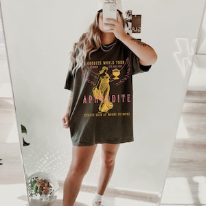 May include: A dark green t-shirt with a graphic print of a woman holding a cup. The text on the shirt reads "Goddess World Tour", "22.4611", "5th Aug 1998", "Aphrodite", and "Tickets sold at Mount Olympus".