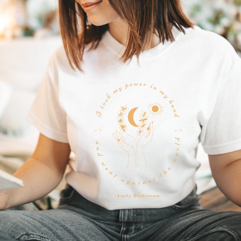 May include: White t-shirt with a gold graphic of hands holding a crescent moon and sun with a floral wreath. The text "I look my power in my hand and went against the world" is printed in gold with the author "Emily Dickinson" below.