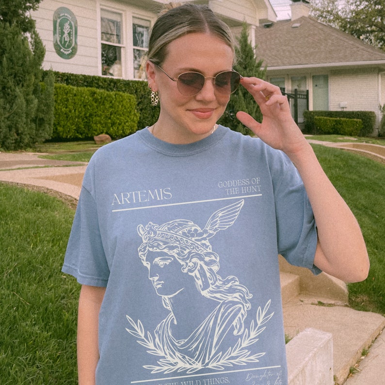 May include: A light blue t-shirt with a white graphic of Artemis, the Greek goddess of the hunt. The graphic includes a bust of Artemis wearing a crown and holding a bow and arrow. The text "ARTEMIS" and "GODDESS OF THE HUNT" is printed in white on the shirt.