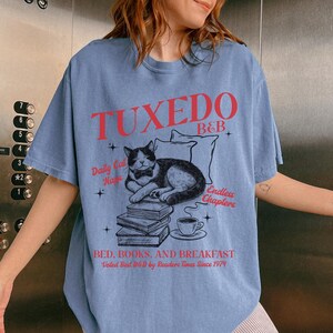 Op de afbeelding: Een lichtblauw T-shirt met een grafisch ontwerp van een kat met een strikje die een boek leest. De tekst op het T-shirt luidt "Tuxedo B&B", "Daily Cat Naps", "Endless Chapters" en "Bed, Books, and Breakfast. Voted Best B&B by Readers Times Since 1974".