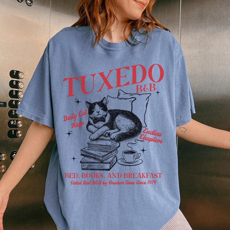 Op de afbeelding: Een lichtblauw T-shirt met een grafisch ontwerp van een kat met een strikje die een boek leest. De tekst op het T-shirt luidt "Tuxedo B&B", "Daily Cat Naps", "Endless Chapters" en "Bed, Books, and Breakfast. Voted Best B&B by Readers Times Since 1974".