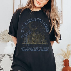 K&ouml;nnte beinhalten: Schwarzes T-Shirt mit einem grafischen Design einer Skyline mit einer zunehmenden Mondsichel und Sternen. Der Text "NYX STARGAZING TOURS" ist kreisf&ouml;rmig um den Mond geschrieben. Der Text "OPEN FROM DUSK TILL DAWN TICKETS FOR SALE AT EREBUS" steht unter dem Grafik.