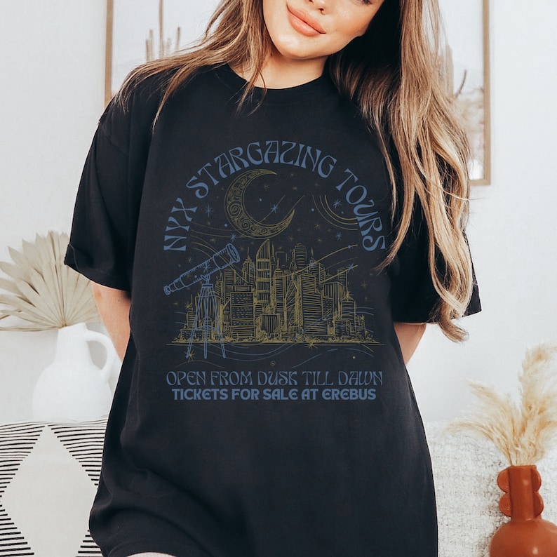 K&ouml;nnte beinhalten: Schwarzes T-Shirt mit einem grafischen Design einer Skyline mit einer zunehmenden Mondsichel und Sternen. Der Text "NYX STARGAZING TOURS" ist kreisf&ouml;rmig um den Mond geschrieben. Der Text "OPEN FROM DUSK TILL DAWN TICKETS FOR SALE AT EREBUS" steht unter dem Grafik.