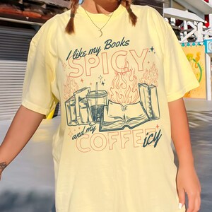 May include: Yellow t-shirt with the text "I like my Books SPICY and my COFFEEicy". The graphic includes illustrations of books, a coffee cup, and flames. The design is in shades of red, blue, and green.