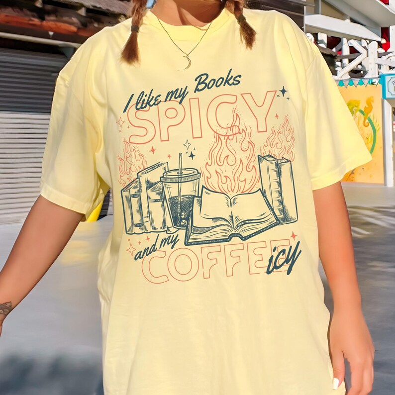 May include: Yellow t-shirt with the text "I like my Books SPICY and my COFFEEicy". The graphic includes illustrations of books, a coffee cup, and flames. The design is in shades of red, blue, and green.