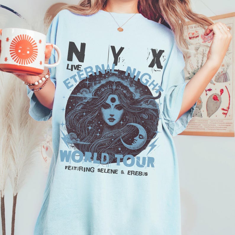 May include: Pale blue t-shirt with a graphic of a woman's face, a crescent moon, and the text "NYX LIVE Eternal Night World Tour Featuring Selene & Erebus." A white and orange mug with a sun design is held in the hand.