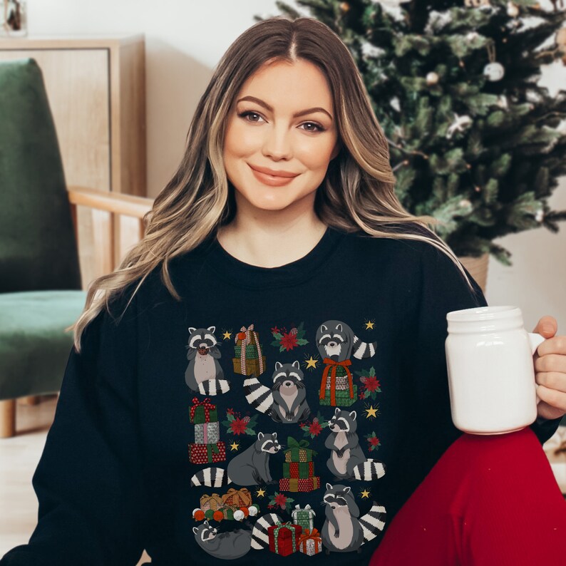 K&ouml;nnte beinhalten: Ein schwarzer Pullover mit einem weihnachtlichen Design, das Waschb&auml;ren zeigt, die Geschenke halten und von Sternen, Stechpalmen und Ornamenten umgeben sind.