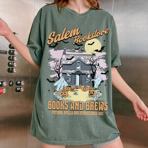 Op de afbeelding: Groen T-shirt met een grafische print van een spookhuis, pompoenen en geesten. De tekst "Salem Bookstore" en "Books and Brews" staat op het T-shirt gedrukt. De tekst "Potions, Spells and Stories Since 1692" staat onder de grafische print.