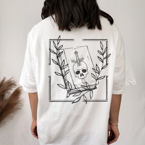 May include: White t-shirt with a black and white graphic print of a book with a skull and a dagger inside. The graphic is framed by a black square with leaves around it.