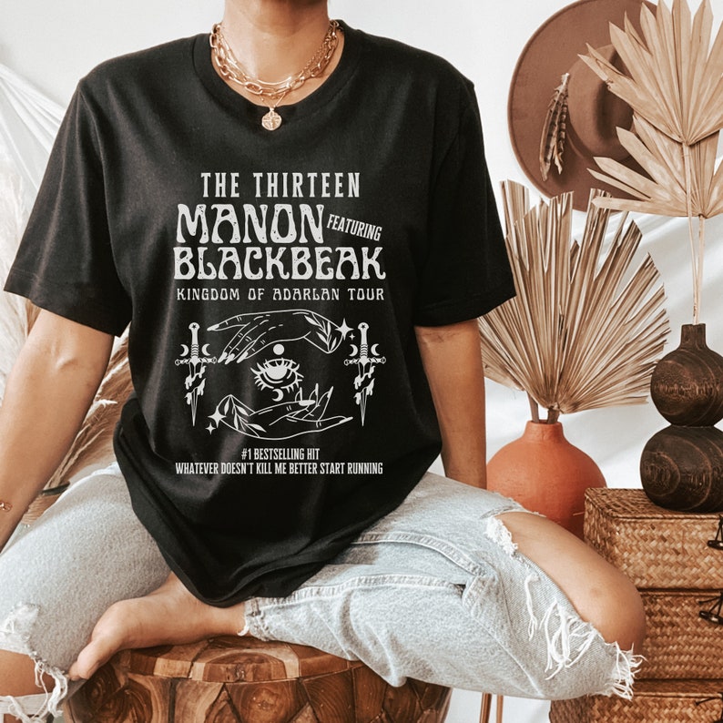 May include: Black t-shirt with white text and graphic. The text reads "The Thirteen Manon Featuring Blackbeak Kingdom of Adarlan Tour #1 Bestselling Hit Whatever Doesn't Kill Me Better Start Running". The graphic is a stylized image of two hands holding a crescent moon and a star with an eye in the centre.