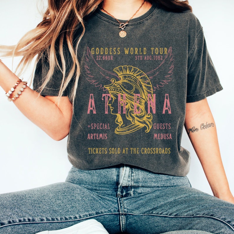 Puede incluir: Una camiseta negra con un gr&aacute;fico de estilo vintage de una mujer con un casco con alas. El texto dice "GODDESS WORLD TOUR", "ATHENA", "22:66NR", "5TH AUG.1982", "+SPECIAL ARTEMIS", "GUESTS MEDUSA", y "TICKETS SOLD AT THE CROSSROADS".